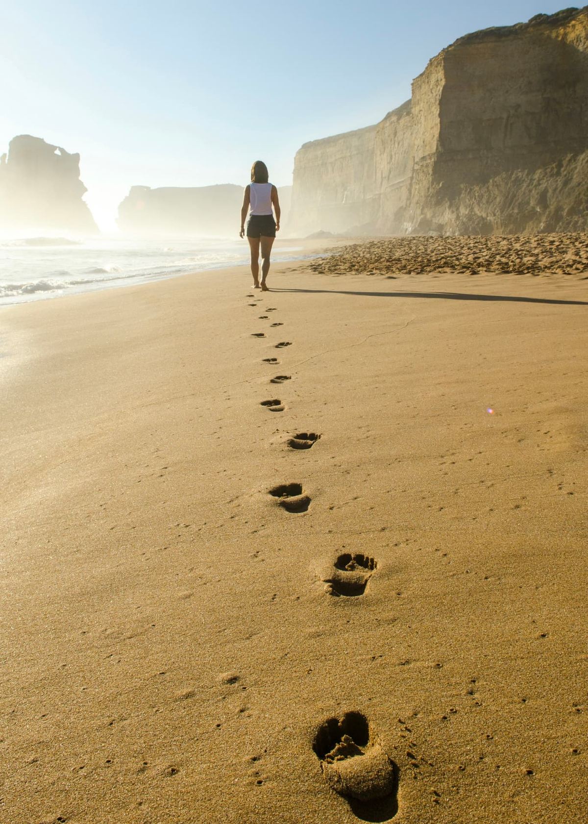 A woman walking barefoot along a wide beach at sunrise, footprints trailing behind her towards distant cliffs